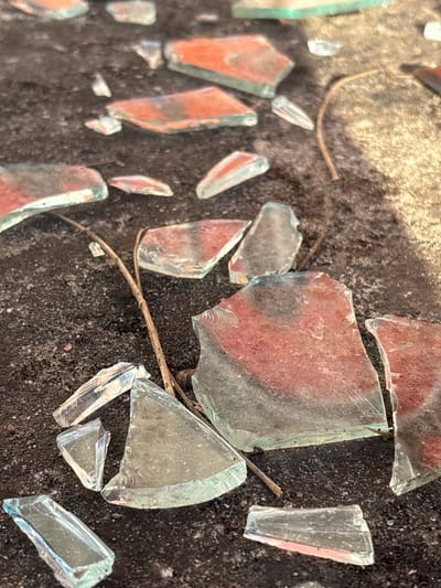 Shattered glass fragments scattered on a dark, rough surface, possibly asphalt or concrete. The glass pieces vary in size and shape, with some exhibiting a reddish tint. A thin, brown twig meanders among the fragments. The background is divided between a dark, textured area and a lighter, blurred patch. The location is stated to be Bratica-Braticë, Montenegro, but this is not visually verifiable. No people, actions, or interactions are visible.