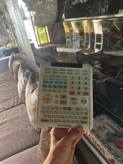 A person is holding a white remote control in their right hand, with their fingers positioned around the bottom half of the device. The remote has a keypad with numbers and letters, along with various function buttons. The background is blurred, but appears to be an indoor setting with furniture that suggests a living room or lounge area. There are elements of decor visible, including what looks like a mirrored surface with a marble-like pattern. The overall lighting is dim, with some light sources contributing to reflections on the surfaces.
There is no information or visual evidence in the image to determine the location within the city of Brca, Montenegro.