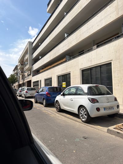This is a medium-shot, eye-level photograph taken from inside a car looking out at a street scene. The image is oriented vertically, as if the camera was held at a 90-degree angle. The main subjects are several cars parked along the side of a street in front of a modern building. The closest car is white, with a license plate that reads "FD-030-VT". Behind it are a blue car and then more white cars. A "A LOUER" (For Rent) sign with a phone number is visible on a window of the building. The building itself is light-colored, with many parallel horizontal concrete beams and narrow windows. The sky above is blue with scattered white clouds, suggesting a clear day. The sun is casting shadows, indicating it is likely daytime. The street is paved with asphalt. The reflection in the side mirror of the car suggests the photographer is wearing a red jacket. The overall scene depicts a typical urban street in France, possibly residential or commercial, on a sunny day.