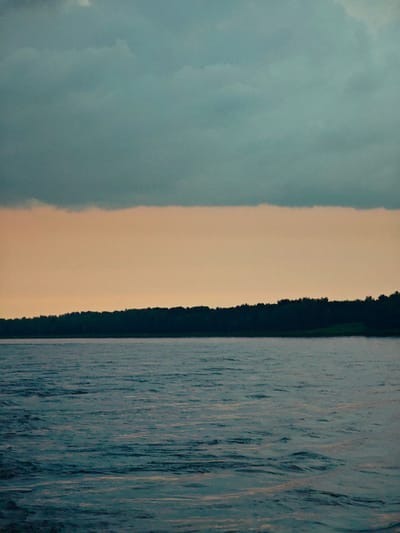 A wide shot captures a body of water under a dramatic sky. The water, a deep, mottled blue, exhibits gentle ripples and reflections. A dark, dense tree line forms a distinct horizon, separating the water from the sky. Above the tree line, a band of pale, peachy-orange sky transitions into a darker, teal-colored sky filled with soft, diffused clouds. The scene evokes a sense of twilight or an approaching storm. The location is the Angara River in Kazachinskoe, Russia.