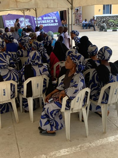 The image shows an outdoor gathering under a large white tent in Ozuri, Nigeria. Numerous individuals, primarily women, are seated on white plastic chairs, many marked with "Aazi." A significant number of the women wear matching blue, white, and black patterned headwraps and flowing garments. In the foreground, a woman wearing this attire is seated on a white plastic chair, facing left with her eyes closed or downcast, and wearing gold hoop earrings and black sandals.
In the background, a large purple banner is displayed, featuring white text that reads "DAM MEDICAL OUTREACH & EMPOWERMENT" and the number "25," likely indicating a foundation or anniversary. Several individuals, including children, are visible standing behind the seated crowd. Further in the background, a building with a light-colored, possibly yellow or cream, facade and a dark, stone-patterned lower section is visible. Other individuals are observed standing on a raised platform or steps in front of this building. The ground is paved, suggesting a courtyard or open area.