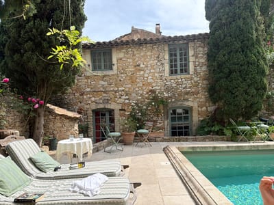 The image depicts a picturesque scene in Saint-Victor-la-Coste, France. A stone building with two windows on the upper level and two doors on the ground level stands as a backdrop. The windows and doors are painted a muted green. The building is flanked by tall, dark green cypress trees on either side. In front of the building is a patio with lounge chairs featuring green-striped cushions and pillows. A small round table with a white tablecloth holds two glasses. Near the center of the image are two small metal chairs positioned around potted plants. To the right, a turquoise swimming pool with visible water reflection borders the scene. A book, sunglasses, and a white cloth rest on the lounge chairs, suggesting a relaxed atmosphere. A partial view of a hand is visible in the lower right corner of the image.