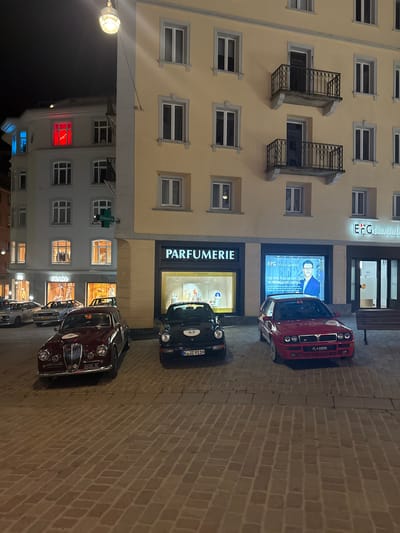 The image depicts a nighttime street scene in Sankt Moritz, Switzerland, specifically on a cobblestone plaza fronting two multi-story buildings. The building on the right features a light yellow facade with multiple windows and two balconies with black wrought-iron railings. Its ground floor houses a "PARFUMERIE" with "ATKINSONS" branding in its display window, and "EFG Private Banking" with a digital display showing a male portrait and German text, alongside a dark entrance and a wooden bench. The building on the left has a white facade with numerous windows; its ground floor displays a "PRADA" storefront and an "ANSELMA" store. A green cross sign labeled "AMAVITA" is affixed to this building. On upper floors of the white building, one window is illuminated in red with visible white text, and another is illuminated in blue.

Three classic or vintage cars are parked prominently on the cobblestone surface. From left to right: a dark burgundy vehicle, possibly a Lancia, with a prominent chrome grille, bearing race number "18" on a white disc and an Italian license plate "2417SY MI"; a dark blue or black sports car, resembling a Porsche 911, with race number "62" on a white disc and a German license plate "K JZ 911H"; and a red rally-style car, likely a Lancia Delta Integrale, with race number "63" on a white disc and a Liechtenstein license plate "FL 20595". Two additional silver vehicles are parked further in the background near the "PRADA" store. The ground consists of light brown rectangular paving stones laid in a basketweave pattern. Overhead, a single streetlamp emits a warm yellow light. No individuals are visible.