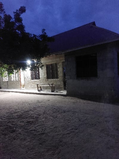The image depicts an outdoor night scene in Mwandege, Tanzania, centered on a single-story concrete block building under a dark blue, cloudy sky. The foreground consists of uneven, dark earth or sand. The building's left section features visible windows with grates or louvers, and an open doorway emitting a bright interior light source. A bare, bright light fixture is mounted on the exterior left wall, illuminating the area directly in front. A rudimentary wooden bench with an A-frame base is positioned on the ground before this illuminated section. The right section of the building appears unfinished or under construction, with dark, empty window openings and concrete block walls. The building is topped with a dark, corrugated metal roof. A large, dark-leafed tree is partially visible on the far left, obscuring a portion of the sky and the leftmost structure. No individuals are present.