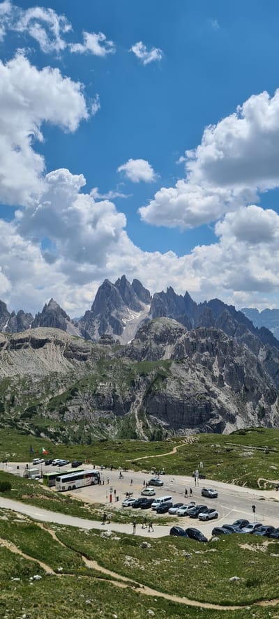 This media file captures a scenic mountain landscape with a parking lot and a bus. The scene is dominated by rugged, gray, rocky mountains that stretch across the frame, with patches of green grass and vegetation clinging to their slopes. A bright blue sky filled with fluffy white clouds provides a dramatic backdrop.

In the foreground and mid-ground, a paved road winds through the terrain, serving as a parking area for numerous cars. Several people are visible walking around the parking lot and along a path that borders the road. A large white and green bus is parked prominently.

The image appears to be taken on a sunny day, suggested by the bright sky and the distinct shadows cast by the mountains and objects. The overall impression is one of a popular tourist destination in a mountainous region, likely during daytime hours. The location cues, such as the Italian flag visible near the bus, combined with the context provided, suggest this is Auronzo di Cadore, Italy. No discernible text is visible in the image.