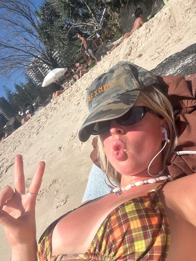 A woman in a bikini top and cap is lying on a sandy beach. The location is determined to be Coolangatta, Australia. The woman is wearing sunglasses, and the cap has orange lettering visible. She is wearing a beaded necklace and appears to have headphones in her ears. She is making a kissing face toward the camera and holding up a hand making a peace sign. In the background, there are people relaxing on the beach and some trees and an umbrella. The sky appears blue.