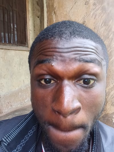 This media file is a close-up, Dutch angle photograph of a person's face. The subject appears to be an adult male with dark skin and short, dark hair. The person is wearing a dark shirt with a patterned trim. The background is a textured, light-colored wall, possibly concrete or stucco, with some darker marks and smudges. To the right of the subject's head, a portion of a barred window is visible. The lighting suggests an indoor setting, possibly during the day. The image is sharp and focused on the subject's face, particularly the eyes, nose, and forehead, which shows visible lines. The overall impression is a candid portrait.