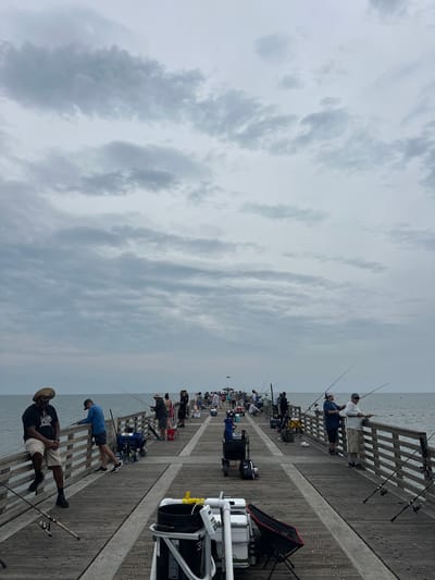 The image depicts a crowded fishing pier at Jacksonville Beach, Florida. Numerous individuals are engaged in fishing, lining both sides of the wooden pier. They are equipped with fishing rods, tackle boxes, and chairs. The sky is overcast with heavy clouds. The pier structure includes wooden planks and railings. The ocean is visible beyond the pier. The scene suggests a popular fishing spot on a cloudy day.