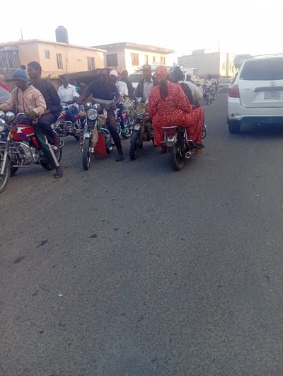 This image captures a bustling street scene in Ozuri, Nigeria, dominated by numerous motorcycles and people, suggesting an urban or semi-urban environment.
The primary subjects are the many people, mostly men, riding or sitting on motorcycles. In the mid-ground, a woman wearing a distinctive red, patterned outfit is a passenger on a motorcycle, facing away from the viewer. Another man in the left foreground wears a plaid shirt and a blue beanie, while a man in a black t-shirt is positioned centrally on his motorcycle.
A key activity is the presence of heavy traffic or congestion. The motorcycles, commonly known as "okadas" in Nigeria, appear to be either waiting or moving slowly. Several motorcycles have multiple occupants, indicating their use as commercial transport. A white Toyota Highlander SUV is visible on the right side of the road, halted or moving slowly.
Notable details include the asphalt road surface, which shows signs of wear. In the background, low-rise buildings with flat roofs, some featuring water tanks, line the street. The lighting suggests it is daytime, with bright but diffused light, indicating either morning or late afternoon. The visible text includes the brand "Bajaj" on the fuel tank of a motorcycle in the left foreground and the Nigerian license plate "FST-92JGZ" on the white SUV. The scene conveys a sense of everyday life and routine activity in a populated area.