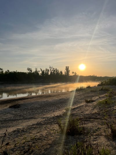 The image captures a sunrise or sunset scene, likely near a body of water. A bright sun is positioned centrally in the frame, radiating light through thin cloud formations. A layer of fog or mist hovers above the water's surface, obscuring the view of the opposite bank, where a dense line of trees is visible. To the right, a sandy bank slopes down to the water's edge, with sparse vegetation clinging to the sand. The scene exhibits calm and natural elements, suggesting a location on the outskirts of Shumerlya, Russia, where rivers and wooded areas may exist. No people or animals are visible.