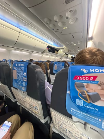 This is a photograph taken from inside an airplane cabin, likely a Boeing 737-600, judging by the visible safety card. The perspective is from a passenger's seat, looking forward and slightly upwards. The overhead compartments are visible, illuminated by blue and white LED lighting. Several passengers are seated in front, their heads and shoulders visible. Some seatbacks have advertising materials, including one with a maze and the text "HOHTOH", and another advertising a "Kreslo-reklayner Rivoa" for 9900 rubles, with a QR code and the website "nonton.ru". A safety-on-board card is also visible on a seatback, detailing emergency procedures. The environment appears to be clean and orderly, typical of a commercial aircraft interior. There is no indication of the city of Сокол, Russia, within the image.