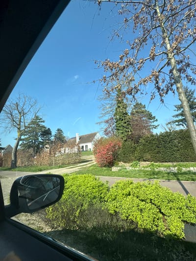 The image captures an outdoor scene from the perspective of a vehicle's passenger side, looking forward and to the right. A dark vehicle frame borders the left and top edges of the photograph. In the lower-left quadrant, a passenger-side rearview mirror is visible, exhibiting a significant crack pattern across its reflective surface.

Immediately outside the vehicle, the foreground features a dense mass of vibrant, bright green, newly-leafed shrubs. A paved path, possibly for pedestrians or bicycles, runs horizontally between these shrubs and a taller, well-maintained, dark green evergreen hedge. Adjacent to the hedge and to the right of a curving gravel driveway, a large bush displays striking bright red-pink foliage or blossoms.

In the midground, the gravel driveway ascends gently towards a two-story white house with a dark, sloped roof and a prominent white chimney. Several windows are visible on the front facade of the house, along with what appears to be a dark-colored door or patio on the lower level. The house is surrounded by a variety of trees; some are tall conifers with green needles, while others are deciduous, currently bare or with sparse, light brown foliage, indicative of early spring.

The background is dominated by a clear, bright blue sky with no visible clouds, suggesting sunny weather. A faint, white linear mark, possibly a contrail or a scratch on the car window, is discernible in the upper sky area. No individuals or discernible activities are present within the scene. The depicted location is Eeklo, Belgium.