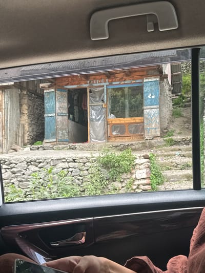 A medium shot captures a weathered, rustic shop with an open entrance, seen through a car window. The shop's facade is a mix of exposed wooden beams and blue painted doors, one of which is ajar, revealing clothing hanging inside. A plastic sheet covers part of the shop's front. The shop is situated on a slightly elevated area, with a stone wall and lush green vegetation in front of it. In the background, a stone wall and more greenery suggest a mountainous or hilly terrain. The foreground shows the interior of a car, with a hand holding a smartphone in the lower left corner and a person's legs covered in fabric on the right. The car's dark interior with a wooden trim and a handle are visible. The location is identified as Kullan, India.