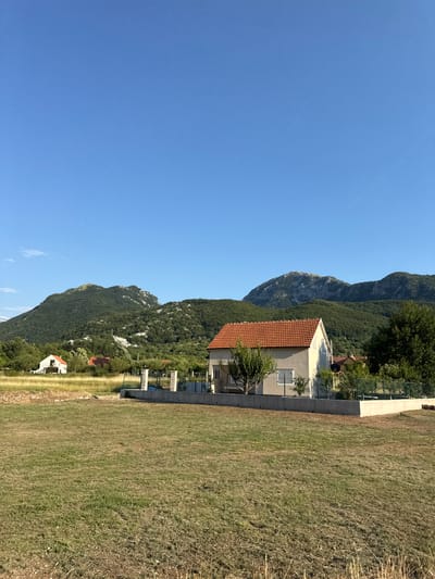 A wide-angle, vertical, outdoor shot shows a residential area in Kopito, Montenegro, with a clear blue sky overhead. In the foreground, a large expanse of dry, patchy grass occupies the majority of the frame. A light-colored house with a red-tiled roof is situated in the mid-ground, right of center, surrounded by a low fence and some greenery. To the left of this house, a smaller white building with a red roof is visible amidst dry grass. In the background, rolling green hills covered in trees and vegetation rise to meet the sky, with rocky peaks visible on the horizon.