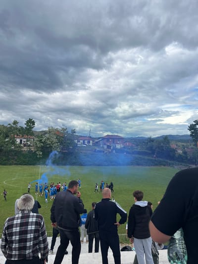 The image depicts a soccer match taking place in Kornitsa, Bulgaria. The scene is observed from an elevated position, possibly a spectator stand, with multiple individuals in the foreground watching the game. Players in blue uniforms are gathered on the green field, with blue smoke emanating from their midst. The background features buildings and a hilly landscape under a cloudy sky. Spectators are visible on the stand, including one wearing a plaid shirt.