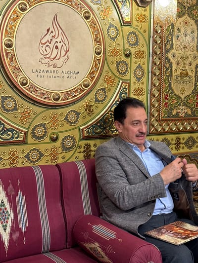 A man sits on a maroon and white striped couch with a matching bolster pillow. He wears a light blue shirt and gray patterned jacket. His head is turned slightly to the right. Overhead is an ornate ceiling with gold, green, and beige geometric patterns. In the center is a circular emblem with Arabic calligraphy and the English text "LAZAWARD ALCHAM For Islamic Arts."