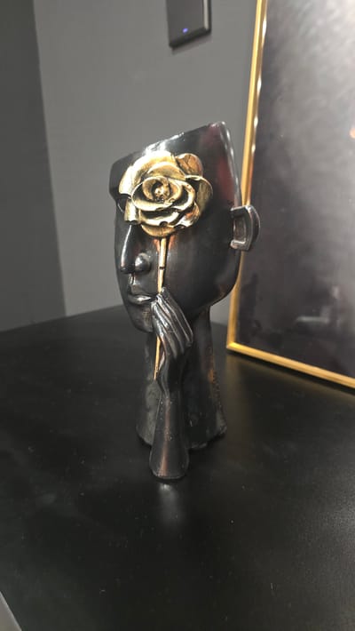 A close-up, eye-level, medium shot captures a dark, abstract sculpture of a human head with a gold rose in its eye socket. The sculpture is positioned on a black, reflective surface, likely a table, in a dimly lit room. The background features a grey wall with a small, black light switch and a portion of a golden framed picture. The sculpture's head is tilted slightly to the left, with a gold rose positioned over the left eye socket. The rose has intricate detailing, with visible petals. The sculpture's left hand is raised to its cheek, appearing to hold the stem of the rose. The right side of the sculpture is partially obscured by the golden frame of the picture in the background. The lighting is subdued, casting shadows on the sculpture and the surface it rests on. The overall impression is one of artistic expression and subtle elegance. The scene does not provide any specific indicators of its location in Trikomo, Northern Cyprus / Cyprus.