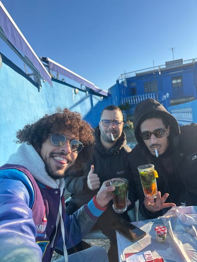 Three men are posing for a selfie while seated at a table outdoors. The man in the foreground has curly hair and sunglasses, and is wearing a blue jacket. The two men behind him are wearing jackets and sunglasses and smoking cigarettes. One is holding a drink with mint in it. A table with drinks, a jar, a phone and cigarettes is in front of them. A blue building is in the background. The sky is clear and blue.