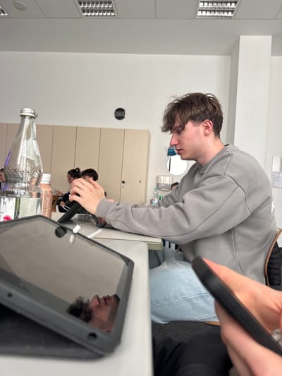 An indoor scene in Bühl, Germany, likely a classroom or study area, depicts multiple individuals seated at light-colored tables under fluorescent ceiling lights. The primary subject, a young male with brown, disheveled hair, wearing a grey sweatshirt and light blue jeans, is seated in profile, looking down and interacting with a small electronic device held in his hands. He is positioned at the right side of the frame. In the immediate foreground, a black-cased tablet lies flat on a table, its screen reflecting the face of another individual (likely the photographer) with dark hair, looking downwards. A white object, possibly a charging cable or stylus, rests on the tablet screen. To the left of the foreground tablet, a clear glass bottle with a silver cap containing effervescent liquid stands next to a light brown bottle. In the background, other individuals are partially visible, seated at tables. A digital clock on a white wall in the background displays "13:53." Behind the seated individuals, light-colored cabinets or lockers are visible, along with a water dispenser unit. A hand holding a dark-colored smartphone is visible in the bottom right corner of the frame. The floor is covered with gray carpeting.