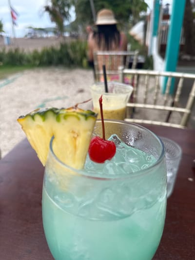 A close-up shot shows a blue cocktail in a glass on a wooden table, garnished with a pineapple wedge and a maraschino cherry. In the background, slightly out of focus, a person wearing a straw hat and a patterned top is visible, along with other drinks and outdoor seating. The scene appears to be at a beachfront establishment in Jacó, Costa Rica, indicated by the sand, vegetation, and beach chairs. The lighting suggests it is daytime, possibly late afternoon.