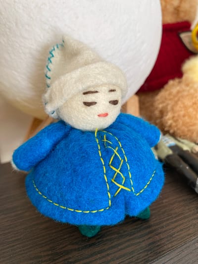 This is a close-up, overhead shot of two small felt toys. On the left is a white, plush toy with simple stitched features: two dark brown eyes and a small pink mouth. It appears to be a bunny or animal of some sort. On the right is a blue, round, plush toy, possibly a ball or a character's head, with yellow stitching outlining a decorative pattern. A small green bud-like shape protrudes from the top right of the blue toy. The toys are positioned so that the white toy's head is nestled against the blue toy's body.

The background is a mix of out-of-focus plush toys and a dark wooden surface. To the left and behind the white toy, a larger white plush surface is visible. Further back, hints of other toys appear, including a red fabric and a fuzzy tan material. The wooden surface, which appears to be a table or floor, has a prominent grain pattern. The lighting suggests an indoor setting, possibly with natural light. There is no discernible text in the image. The scene is static, with no activity taking place beyond the arrangement of the toys.
