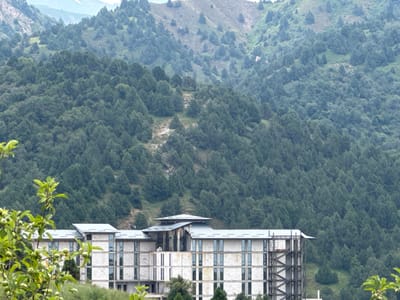 The image depicts a multi-story building under construction, with a light-colored facade and numerous windows. The building is situated in a mountainous, forested area. The foreground features a branch of a tree with green leaves and small green fruits, partially obscuring the lower section of the building. In the background, a dense forest covers the slopes of rolling hills, with some sparser vegetation and rocky patches visible higher up. The sky is overcast and light-colored, suggesting a diffused light condition. The overall scene conveys a sense of a remote, natural setting with ongoing construction activity. This scene is located in the city of Uchterek, Uzbekistan.