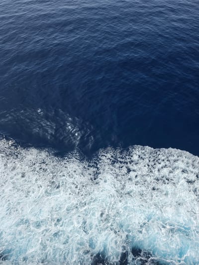 This is a close-up, high-angle shot of the ocean's surface. The top two-thirds of the image are dominated by deep blue water with subtle ripples and surface texture. The bottom third transitions into a churning mass of white foam and lighter blue water, indicative of a wake. The foam is dense and textured, with many small, irregular bubbles and streaks of lighter blue water interspersed. There are no people, objects, or identifiable landmarks visible in this abstract depiction of ocean water. The scene is one of movement and texture within the marine environment.