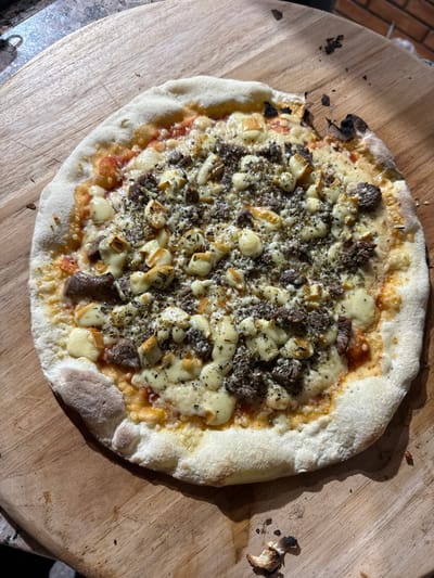 A freshly baked, circular pizza is centrally positioned on a light-colored wooden pizza peel. The pizza, approximately 14 inches in diameter, features a well-risen crust with localized browning and charring. Its surface is covered with a base layer of tomato sauce, melted white cheese, numerous distinct small cubes of pale yellow cheese, and finely minced, dark brown cooked meat. A substantial amount of dried green herbs is distributed over the toppings. Scattered around the pizza on the wooden surface are small, dark, charred debris fragments. In the upper right background, a reddish-brown brick wall is partially visible. The scene occurs in a domestic or casual culinary setting within Porto Alegre, Brazil, under natural light from the left, casting a pronounced shadow of the pizza onto the board. No individuals are present in the frame.