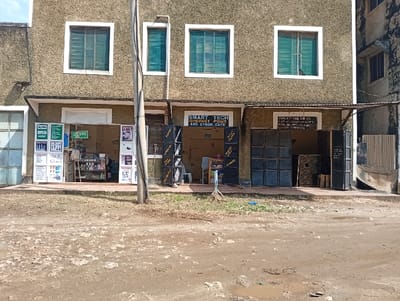 This image depicts the exterior of a two-story commercial building located in Shella ward, Kenya. The building features a textured, light brown or beige facade with four rectangular windows on the upper floor, each fitted with green blinds or shutters and white frames.
The ground floor houses multiple businesses. On the left, an open shop is visible, displaying white advertising boards for "M-PESA Agent" and various products, including cosmetics and mobile services. Inside, shelves are stocked with merchandise. In the center, a business identified by a sign as "SMART TECH INTERNET POINT AND CYBER CAFE" has an open entrance flanked by multiple large, black sound system speakers. A table and chairs are discernible within its interior. To the right, another establishment bears a sign reading "MALIZA FOOLS TRADING" with phone numbers; its entrance is partially closed by dark, multi-panelled doors. An adjacent, darker building segment is visible on the far right.
In the immediate foreground, an unpaved dirt road with an uneven surface and small depressions is present. Patches of sparse, dry vegetation are scattered along the roadside directly in front of the building. A tall, light-colored utility pole stands centrally in the foreground, slightly to the left of the "Smart Tech" business, with wires extending upwards. No individuals, actions, or interactions are visible within the scene.