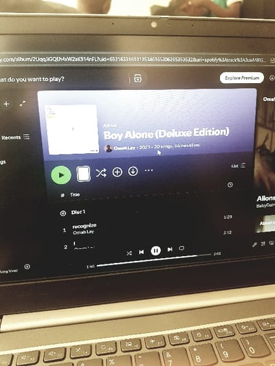 This image captures a close-up view of a laptop screen and a portion of its keyboard, located in the city of Jama A, Nigeria. The laptop display prominently features the Spotify application interface. The main subject on the screen is the album "Boy Alone (Deluxe Edition)" by Omah Lay, indicated as released in 2023, comprising 20 songs and having a total duration of 64 minutes 41 seconds. A mouse cursor is visible hovering over the album's duration information. The tracklist for "Disc 1" is partially displayed, showing "1. recognize" and "2. I". A playback progress bar indicates 1 minute 40 seconds of elapsed time for the current track. The laptop's webcam is visible at the top center of the screen bezel. The keyboard, showing function keys and number keys, is in the lower foreground. In the blurry background, a partial human hand is visible on the right, and a indistinct figure wearing a light-colored top is also present.