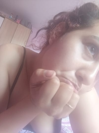 A person is depicted in a close-up, eye-level view, lying on their side and looking upwards and to the right of the frame. The individual has dark, curly hair, visible at the top and left of the head. A light-colored eye, possibly blue or grey, is prominent on the right side of the face. One hand is raised to the face, covering the chin and lower lip. A simple, thin gold-colored band ring is worn on the index finger of the raised hand. A dark-colored bra strap is visible over the left shoulder. The skin tone appears light to medium. The background features a light pink or lavender-colored wall. A light brown wooden cabinet or wardrobe with two doors is situated against the wall on the left side of the frame. Various objects are placed on top of the cabinet, including bottles (one appearing to be an aerosol can) and what seems to be a pink bag or item. A patterned textile, possibly bedding or a cushion, with white and dark purple/pink geometric designs is visible at the bottom right. The specific location within the city of Vinitsa, Bulgaria, cannot be determined from the visual information provided in the image.