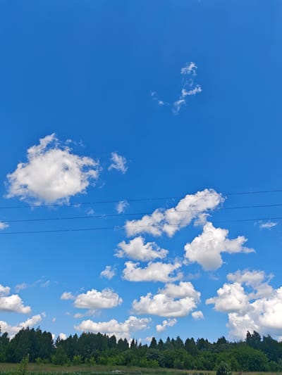The image depicts a bright, clear sky with scattered white cumulus clouds. Three thin, dark electrical wires stretch horizontally across the middle of the frame. Below the sky and wires, a dense line of green trees forms the horizon, suggesting a natural landscape. The ground in the foreground appears to be a mix of green grass and possibly some distant fields. The scene is consistent with an outdoor view, likely in a rural or semi-rural area. While the image is visually rich, it lacks specific details to pinpoint a location within the city of Кожласола, Russia, beyond it being an outdoor scene in that general region. There are no people, animals, or specific man-made structures visible that would offer further clues to a precise location.