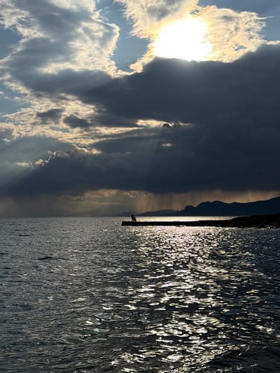 The image shows a body of water with reflections of sunlight, indicating waves or ripples on the surface. On the left side of the frame, there's a sky with clouds, and the sun is visible, partially obscured by the clouds. A dark, mountainous shape is visible in the middle distance. A thin vertical line, likely a pier or similar structure, is located between the water and the land. A silhouette of what appears to be a person is visible on the pier. The location is Novyi Svet, Ukraine.