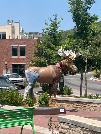 This is a daytime, eye-level, outdoor, full-shot photograph of a large, brown moose sculpture located in a public space in Park City, United States of America. The sculpture is adorned with a yellow cowboy hat, a red scarf around its neck, denim jeans on its hindquarters, and brown boots on its hind legs. The moose is positioned in front of a brick building with multiple windows. To the left of the sculpture, a dark-colored vehicle and a white pickup truck are visible. The background features green trees and a hillside with houses. In the foreground, there is a green metal bench and a low stone wall. The ground is paved with red bricks.
