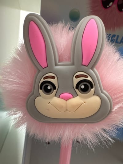 A close-up, eye-level, vertical shot shows a pink, fluffy, pom-pom-like object with a gray cartoon bunny face attached to the front. The bunny has large, pink inner ears, large gray outer ears, brown eyebrows, black eyes with white pupils, and a small pink nose. Its mouth is represented by a simple curved line. The background is blurred and appears to be indoors, with a partially visible blue and white object with text that reads "OLA" on the right side, and a white surface on the left. The specific location in Keila, Estonia, cannot be determined from the visual information provided.