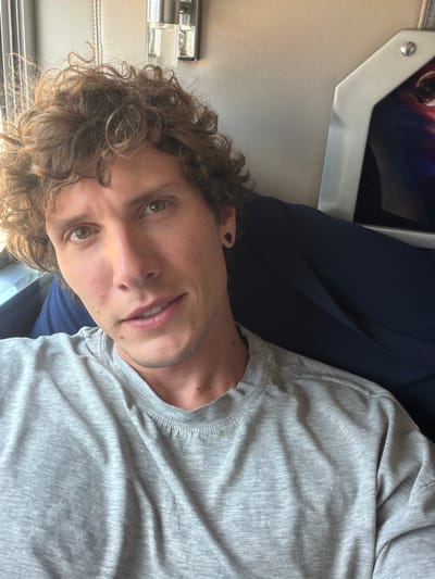 A man with curly brown hair and green eyes is shown from the chest up, looking directly at the camera. He is wearing a grey t-shirt. He has a small earring in his left ear. He appears to be sitting on a train, with a dark blue seat behind him and a silver panel with a picture of a planet on the right side of the frame. The lighting suggests it is daytime. The man's skin has a slight tan and he has a few moles visible on his face. His lips are slightly parted, revealing his teeth.