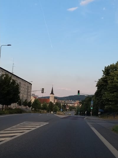 The image shows a street scene in Strakonice, Czechia, looking towards a church steeple and a town in the distance. The foreground features an asphalt road with a pedestrian crossing marked by white stripes. On the left, a multi-story building with many windows is visible, along with trees and a lamppost. Traffic lights are positioned on poles along the road, with one showing a green light. The sky is blue with a thin white contrail from an aircraft. The overall impression is a quiet urban environment during daylight.