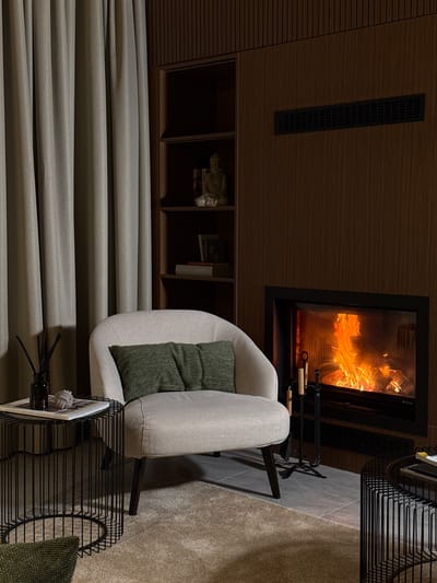 The image depicts a cozy living room scene in Bakovići, Montenegro. A plush, light-colored armchair with two green throw pillows sits on a textured beige carpet. To the left of the armchair is a black wireframe side table with a dark glass diffuser bottle and a book. In the background, a fireplace with a roaring fire is set into a dark wood-paneled wall. Above the fireplace is a horizontal vent. To the left of the fireplace is a dark wood shelving unit containing a small Buddha statue and books. A tall, light gray curtain hangs on the far left side of the image. The overall ambiance is warm and inviting.