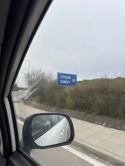 The image depicts a roadside scene captured from within a vehicle, likely located near the Lesovo border crossing in Bulgaria, indicating proximity to Turkey. A blue rectangular road sign, prominently positioned on an embankment to the right, displays "ТУРЦИЯ" (TURKEY) above "TURKEY," followed by "0.5," signifying 0.5 kilometers to the Turkish border. The sky is overcast and uniformly grey. The foreground shows a portion of the vehicle's white interior door frame and a side mirror. The mirror's reflection reveals the occupant of the vehicle, wearing a light green shirt. To the left, a paved road is visible, curving slightly, with several bare-branched trees and streetlights in the distance. The area adjacent to the road on the right features a concrete sidewalk or shoulder, bordering a gentle slope covered with dry bushes and some green vegetation.