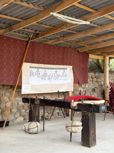 This image captures an outdoor workshop or exhibition space in the city of Кызыл-Туу, Kyrgyzstan. The scene is set under a rustic wooden roof supported by beams, with corrugated roofing material overhead. A patterned red textile hangs as a backdrop.
Dominating the center of the image is a large, hand-drawn diagram or chart displayed on a piece of paper, possibly a family tree or organizational chart, with rows of small, house-like icons connected by lines. The text above and below the chart appears to be in a Cyrillic-based script.
In front of the chart is a sturdy, heavy wooden workbench. This workbench is supported by substantial wooden legs and appears to be a functional piece of equipment for craft or trade. Suspended from the workbench are two large, round stones, each wrapped with wire or rope for stabilization and potentially for use as weights or counterbalances. A long, wooden pole, possibly a tool or staff, leans against the left side of the workbench.
The environment suggests a rural or traditional setting, with a stone wall visible behind the workbench and some greenery in the background. The ground is concrete. Red materials are also visible on the workbench, suggesting ongoing work or display. There are no people visible in the frame.