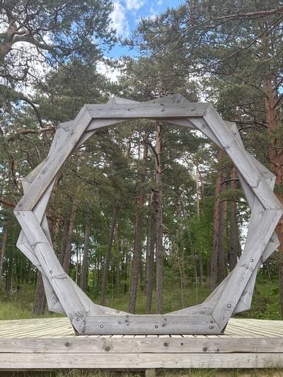The image displays a large, weathered wooden archway framing a view of a pine forest under a clear sky. The archway, constructed from angular segments of light grey wood, forms an irregular polygonal shape, possibly an octagon. Each segment is visibly secured by screws or bolts, highlighting the structure's construction.
The archway is anchored into a wooden platform or boardwalk visible on the right side of the frame, with some green grass and dry vegetation at its base. Through the opening of the archway and beyond its edges, numerous pine trees with their tall, straight trunks and dense green canopies fill the background. Patches of bright blue sky with sparse white clouds are visible overhead through the tree branches.
The scene suggests a daytime setting with pleasant weather. There are no people present or any specific activities taking place, creating a tranquil and natural atmosphere. The wooden structure appears to be an outdoor feature, possibly a scenic lookout or an art installation, within a forest environment. The location context indicates this is in Saulkrasti, Latvia. No visible text is present in the image.