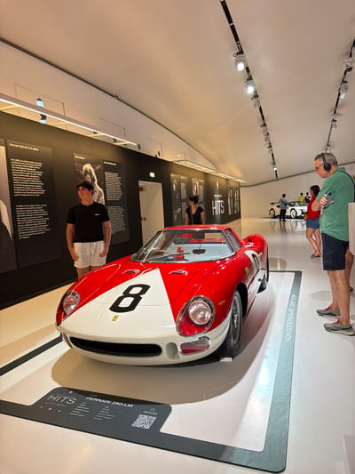 This image was taken at the Ferrari Museum in Modena, Italy. The main subject is a vintage red and white Ferrari 250 LM race car, numbered "8", displayed on a white platform in a museum exhibition space. The car is positioned prominently in the foreground.
Several people are visible in the background. To the left, a young man wearing a black t-shirt and white shorts stands near an information panel about the Ferrari 250 GT 2+2 1960. To the right, an adult male wearing headphones and a green shirt is holding a smartphone, looking towards the display. Further in the background, other museum visitors are observing the exhibits, including another white Ferrari vehicle.
The museum environment features a clean, minimalist design with white walls and flooring. The ceiling has a curved design with track lighting. Information panels with text and images are displayed on the walls, providing context about the exhibits. A sign on the floor near the red car reads "NON OLTREPASSARE - DON'T STEP" indicating a boundary. A display plaque in front of the Ferrari 250 LM identifies it as a "HITS" exhibit featuring the "FERRARI 250 LM".