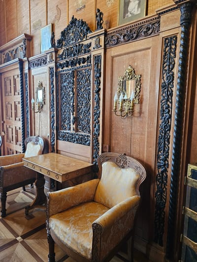 A view of a room in Алупкинский городской совет, Ukraine, featuring an armchair next to a table. The room is characterized by its ornately carved wooden wall panels, embellished with floral motifs and scrollwork. On the walls are sconces and framed photographs. The chair and table are adjacent, with the chair upholstered in a floral-patterned fabric. The floor is patterned with wood.
