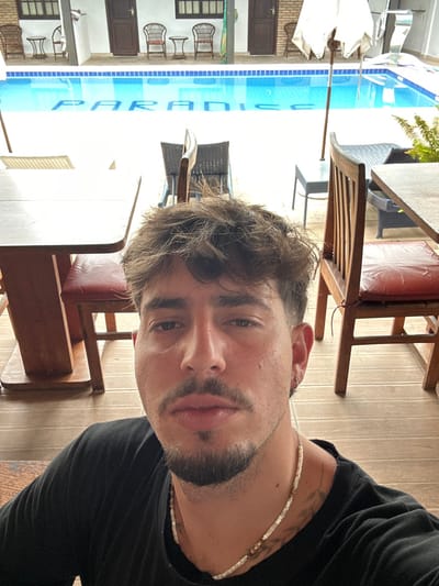 A young adult male, approximately 20-35 years old, is centered in the foreground of a selfie, taken at a resort or pousada in Lauro De Freitas, Brazil. He has short, dark brown wavy hair, a mustache, and a beard. He is wearing a black t-shirt and a light-colored beaded necklace. A silver stud earring is visible in his left ear. He maintains a neutral expression, looking directly at the camera.
The background reveals an outdoor leisure area. Directly behind the subject, a bright blue swimming pool is visible, with the word "PARADISE" spelled in white letters on its floor. Flanking the subject are parts of two wooden dining tables with red-cushioned chairs. Across the pool, a white two-story building features multiple guest rooms, each equipped with a dark brown wooden door and a rectangular window with dark frames. Several wicker chairs and small tables are arranged on the paved deck surrounding the pool. A white market umbrella stands on the right side of the pool, shading a dark grey square table. Green foliage is partially visible on the far right edge of the frame.