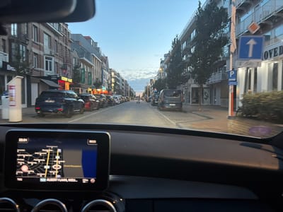 This media file depicts a street scene in Nieuwpoort, Belgium, viewed from the interior of a car. The time of day appears to be late afternoon or early evening, as indicated by the soft, warm lighting and the clear sky with a few wispy clouds. The street is lined with buildings, some of which appear to be shops or residences with multiple stories. Visible businesses include a "Golden Palace" casino and a "Carrefour express" grocery store.

Several cars are parked along the street, and others are driving. A dark blue SUV is prominent in the foreground. The dashboard of the car shows a navigation system displaying a map with the text "ALBERT I LAAN, NIEU..." which strongly suggests the location is Albert I Laan in Nieuwpoort. Road signs indicate "P" for parking and an upward arrow with a bicycle symbol. The overall atmosphere is calm and ordinary, suggesting a typical urban or coastal town setting during a pleasant time of day. There are no distinct individuals clearly visible as subjects, but the presence of parked and moving vehicles implies normal activity within the town.