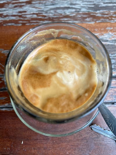 A close-up, overhead shot captures a glass of espresso with crema, sitting on a weathered wooden table. The coffee's surface is a swirling mix of golden-brown crema. A fragment of a metallic object, possibly a spoon or fork, is visible to the right of the glass. The wooden table exhibits a distressed texture with visible grain and patches of lighter-colored wear. The location is identified as Pantegajah, Indonesia. There are no people or explicit actions depicted.