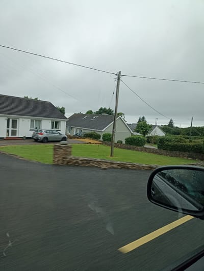 The image captures a view from a car, looking out at a residential street in Dore, Ireland, under an overcast sky. In the foreground, the wet asphalt road is visible, with a distinct yellow line marking its edge. A car's side mirror, reflecting the grey sky and some trees, is positioned in the lower right corner.

Further into the scene, a neatly maintained lawn slopes gently upwards, bordered by a low, rough-hewn stone wall. Beyond the wall, several houses are visible. To the left, a white bungalow with a dark tiled roof features large windows and a front door. A silver hatchback car is parked in the driveway.

To the right of this first house, another bungalow with a similar dark tiled roof and dormer windows is situated. A line of mature green hedges runs along the front of this property and continues to the right, obscuring the bases of the houses behind them. In the background, more houses and dense greenery, including trees, are scattered across the landscape. Power lines and a wooden utility pole are present, crossing the sky. The overall impression is one of a quiet, suburban or semi-rural setting on a damp, cloudy day.
