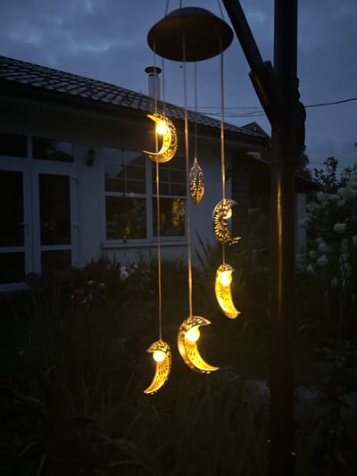 In the twilight hour, a string of crescent moon-shaped solar lanterns hang from a dark pole in the backyard of a house in Romashkino, Russia. The lanterns, each with a warm, glowing bulb inside, are intricately cut with decorative patterns. The house, partially visible in the background, features a dark shingled roof and white-framed windows. Lush green foliage and flowers, some appearing to be white and pink roses, surround the base of the house, contrasting with the darkening sky. The scene is tranquil, illuminated by the gentle glow of the lanterns against the dusky blue of the evening sky.
