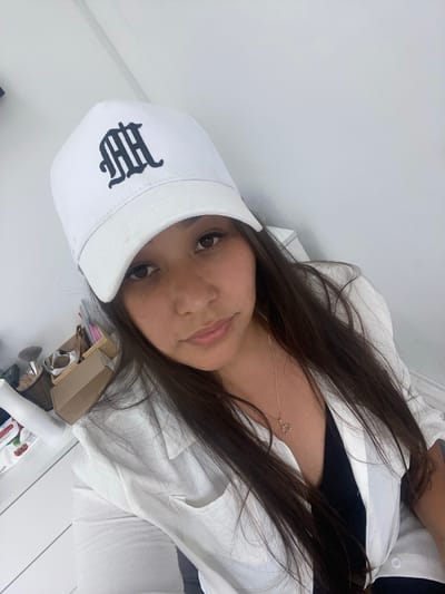 A young woman with long, dark hair and brown eyes is wearing a white baseball cap with a black gothic-style "MH" logo. She has on a white unbuttoned shirt over a black top and a delicate gold necklace with a rose pendant. The background is a plain white wall. To the left of the woman, there is a white dresser or table with various items, including a white spray bottle, a pack of wipes with strawberries on it, a wicker basket filled with makeup brushes, and a cardboard box. The lighting suggests an indoor environment, possibly a bedroom or dressing area. There is no indication of the city of Akyar, Russia, within the image itself.