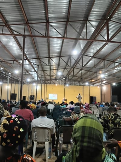 A large indoor gathering takes place under a metal-framed structure with corrugated walls. Rows of individuals sit in chairs, seemingly focused on a central stage area with a screen. The crowd features diverse attire, including patterned fabrics and head coverings. A few individuals stand at the back, observing the event. Overhead lighting illuminates the interior space, casting shadows across the room.
